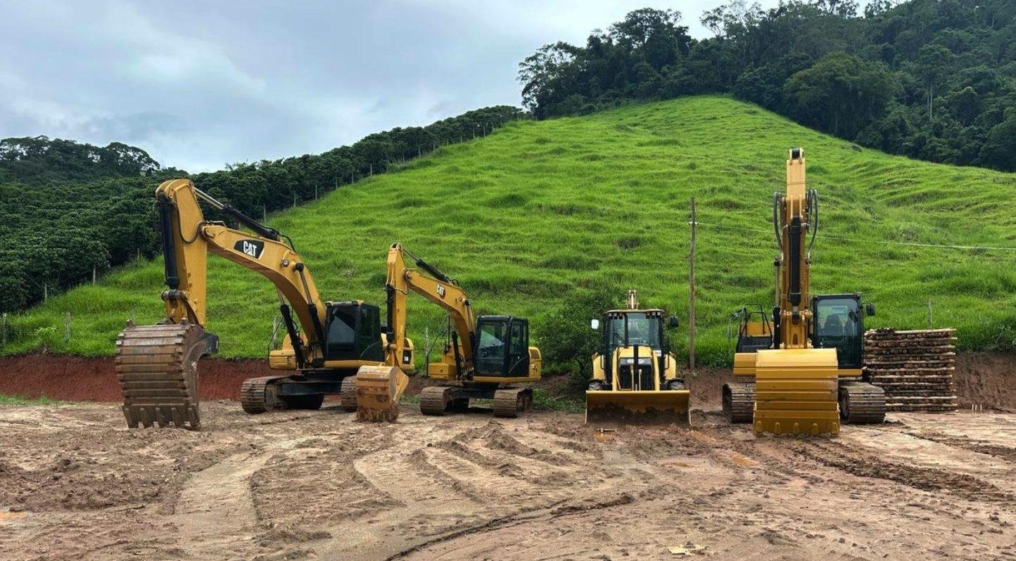 Escavadeiras e retroescavadeira Caterpillar da Escave alinhadas em frente de obra, com morro verde ao fundo